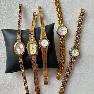 May include: Five gold-tone wristwatches with metal link bands. Three watches feature round or rectangular faces with white dials, while two have intricate patterned bands. The watches are displayed on a black cushion, showcasing various link styles, metallic finishes, and dial designs including Roman numerals and standard hour markers.