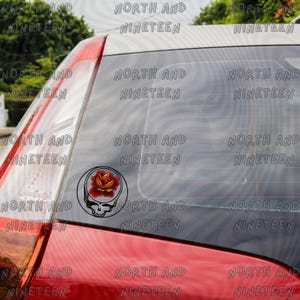 May include: A black and white decal featuring a skull with a rose on a car window. The rose is red and orange. The decal is circular and has a black outline. The text "NORTH AND NINETEEN" is repeated around the window.