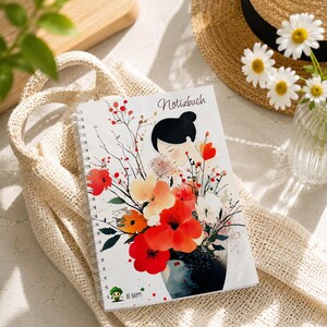Könnte beinhalten: Ein spiralgebundenes Notizbuch mit weißem Einband, das eine Aquarellillustration einer Person mit dunklem Haar zeigt, umgeben von roten, orangefarbenen und weißen Blumen. Oben steht der Text Notizbuch und unten eine kleine grüne Figur mit dem Text BE HAPPY.