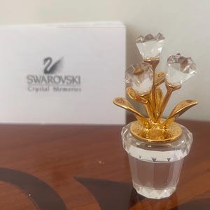 Op de afbeelding: Een Swarovski kristallen bloemstuk in een heldere, facetgeslepen pot met goudkleurige accenten. Het arrangement heeft drie heldere kristallen bloemen met gouden stelen en bladeren. De Swarovski doos staat op de achtergrond.