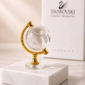 Peut inclure: Un globe terrestre en cristal transparent, monté sur un support en métal doré, est posé sur un bloc de marbre blanc. Le globe est facetté et présente les continents. Une boîte blanche avec le logo Swarovski est en arrière-plan.