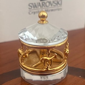 Può includere: Figurina carosello in cristallo con dettagli dorati. La figurina presenta una cupola trasparente e sfaccettata e una base con cavalli dorati. Il carosello è un oggetto decorativo, probabilmente da esposizione.