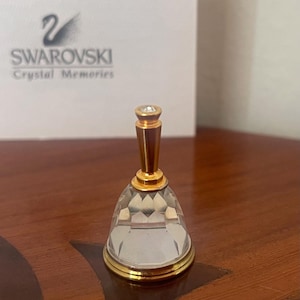 Könnte beinhalten: Eine transparente Kristallglocke mit einem goldfarbenen Griff und Sockel. Die Glocke hat ein facettiertes Design und einen kleinen, klaren Edelstein an der Spitze. Die Glocke steht auf einer Holzoberfläche. Auf der Schachtel hinter der Glocke steht "SWAROVSKI Crystal Memories".