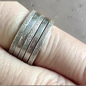 May include: A set of four silver rings stacked on a finger. Two rings have a textured surface, and two have a smooth, polished finish. The word "PANCOADS" is engraved on two of the rings.