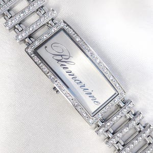 Könnte beinhalten: Eine silberfarbene Armbanduhr mit rechteckigem Zifferblatt und einem Armband, das mit glitzernden Kristallen besetzt ist. Auf dem Zifferblatt steht das eingravierte Wort "Blumarine". Das Armband besteht aus silbernen Gliedern.