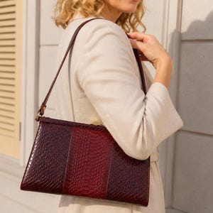 Könnte beinhalten: Eine weinrote Handtasche mit Schlangenmuster. Die rechteckige Tasche hat einen langen, dünnen Riemen und einen goldfarbenen Verschluss. Die Tasche wird von einer Person getragen, die eine cremefarbene Jacke trägt.