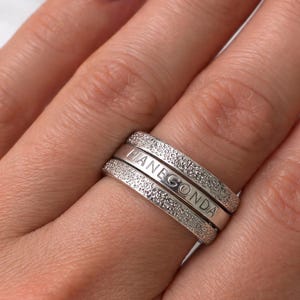 Könnte beinhalten: Drei silberne Ringe, die auf einem Finger gestapelt sind. Der mittlere Ring trägt die Gravur "PIANEGONDA". Die oberen und unteren Ringe sind mit kleinen, funkelnden Steinen besetzt. Die Fingernägel sind tiefrot lackiert.