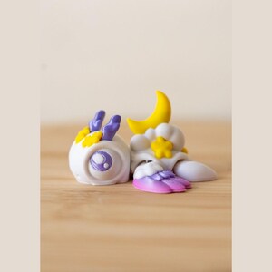 May include: A small white figurine featuring a rounded head with purple antennae and a large purple eye. The body is adorned with a yellow crescent moon and a yellow star, accompanied by a wing-like appendage that transitions from purple to pink.