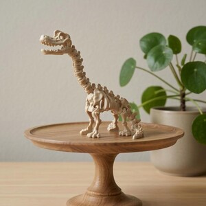 Puede incluir: Una figura de esqueleto de dinosaurio beige sobre un soporte de pastel de madera. El dinosaurio tiene un cuello y una cola largos, con una estructura ósea detallada. El soporte para pasteles es redondo con una base de pedestal. Una planta en maceta está en el fondo.