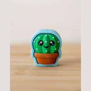 Pode incluir: Uma pequena figura tridimensional de um cacto verde com um rosto sorridente, grandes olhos pretos e pontos brancos, plantado numa base castanha. A figura está inserida numa moldura arredondada de cor azul-clara com textura visível.