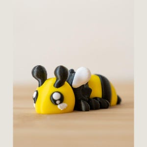 May include: A small, three-dimensional printed bee figurine featuring a bright yellow head and body with black stripes. The bee has two black antennae, large black eyes with white highlights, and a single visible white wing on its side.