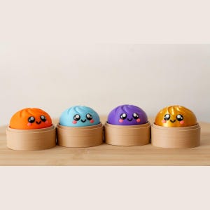 Könnte beinhalten: Vier abgerundete, kuppelförmige Figuren in Orange, Hellblau, Lila und Metallic-Gold, die jeweils ein lächelndes Gesicht mit großen schwarzen Augen und rosa Wangen aufweisen. Jede Figur ruht in einem kleinen, runden, hellen Holzbehälter.