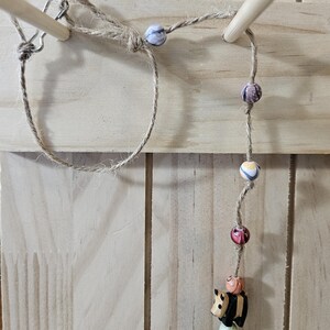 May include: A handmade wooden toy with a panda bear figure and colorful beads strung on a natural fiber rope. The panda is black and white, and the beads are various colors and patterns. The toy is suspended between two wooden dowels.