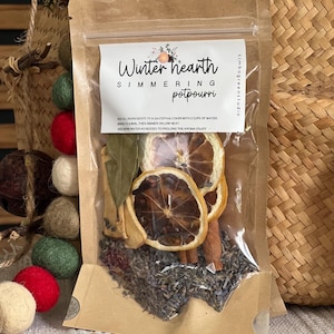May include: A clear, sealed pouch of "Winter Hearth" simmering potpourri. The pouch contains dried orange slices, cinnamon sticks, lavender, and bay leaves. The label includes instructions for use. The background includes decorative felt balls and a woven basket.