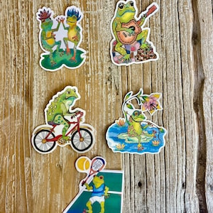 May include: A collection of colorful frog stickers on a wooden surface. The stickers depict frogs engaged in various activities, including playing guitar, riding a bicycle, playing tennis, and dancing. The frogs are in various poses and settings.
