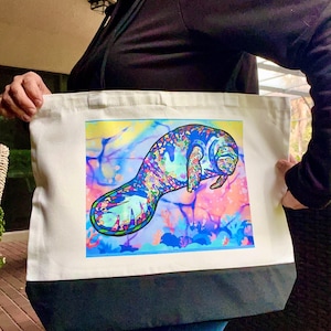 May include: A white tote bag with a black bottom panel. The bag features a colorful illustration of a manatee in shades of blue, purple, and green against a vibrant background of pink, yellow, and blue.