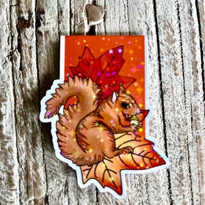 May include: A sticker featuring a brown squirrel holding a nut, set against a backdrop of fall leaves. The squirrel has a fluffy tail and is surrounded by orange and red leaves. The sticker has a holographic, glittery effect.