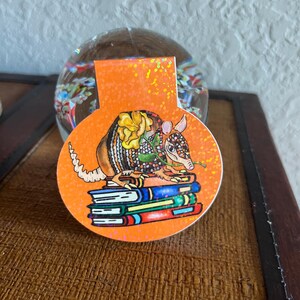 May include: A round, orange bookmark with glitter, depicting a cartoon armadillo on a pile of books. The armadillo has a brown and tan shell and holds a sprig. The books are colourful. The bookmark has a rectangular tab.