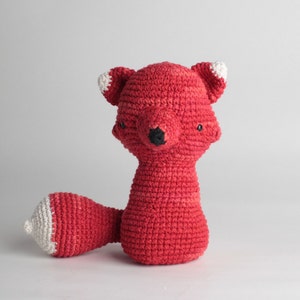 Puede incluir: Un juguete de peluche de zorro rojo tejido a crochet con detalles blancos en las orejas y la cola. El zorro tiene ojos negros y una nariz negra.