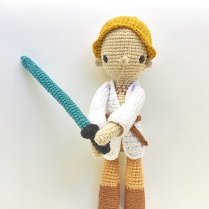 Puede incluir: Una figura de juguete de crochet de un personaje de Star Wars, que lleva una túnica blanca, pantalones marrones y un sombrero amarillo. La figura sostiene una espada láser azul.