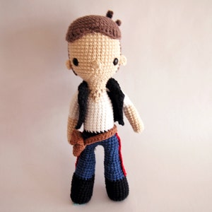 Puede incluir: Un juguete de peluche tejido a crochet de un personaje que lleva un chaleco marrón, una camisa blanca, pantalones vaqueros azules y un cinturón marrón. El juguete tiene el pelo marrón y una cara marrón con ojos negros.