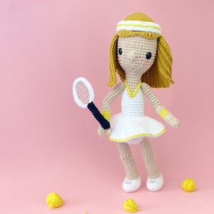 Puede incluir: Una muñeca de crochet vestida con un vestido de tenis blanco con ribetes amarillos, una diadema amarilla y sosteniendo una raqueta de tenis negra y blanca. La muñeca está de pie sobre un fondo rosa con ovillos de lana amarillos esparcidos alrededor.