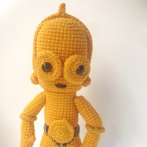 以下が含まれることがあります： スターウォーズの黄色いクロシェのC-3POキャラクター。キャラクターは黒い目と黒い口をしています。キャラクターはカラフルなベルトをしています。