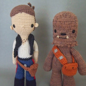 Puede incluir: Dos personajes de Star Wars hechos a crochet, Han Solo y Chewbacca, están de pie uno al lado del otro. Han Solo lleva un chaleco marrón, pantalones azules y un cinturón marrón. Chewbacca es marrón con un bandolera de color beige y un bolso marrón.