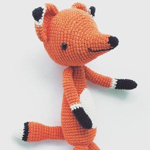 Puede incluir: Un juguete de zorro de crochet con pelaje naranja, ojos negros y detalles en blanco y negro en las patas y la cola.
