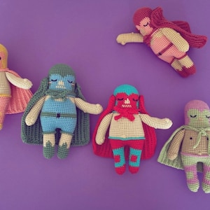 May include: A collection of handmade crochet superhero dolls in various colors, including pink, blue, red, and green. Each doll has a mask and cape, with a unique color scheme. The dolls are arranged on a purple background.