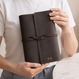 Könnte beinhalten: Ein dunkelbraunes Lederjournal mit einem Wickelverschluss aus Lederband. Auf dem vorderen Einband sind in der unteren rechten Ecke die Initialen V.S. eingeprägt. Das Journal hat sichtbare Nähte an den Kanten und eine glatte, matte Oberfläche.