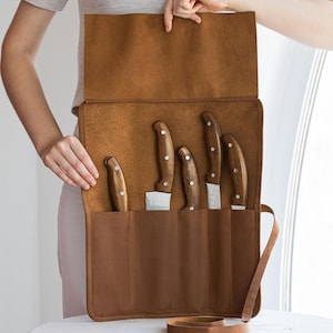Puede incluir: Un estuche de cuchillos de cuero marrón con cinco cuchillos. Los cuchillos tienen mangos de madera y hojas plateadas. El estuche está abierto, mostrando los cuchillos en ranuras individuales. Se ve una correa de cuero marrón.