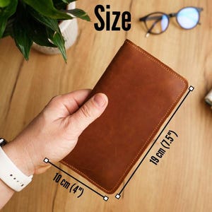 May include: A brown leather wallet, measuring 10 cm wide and 19 cm tall, is held in a hand. The wallet has a stitched border and a smooth surface. The word "Size" is visible in the image.