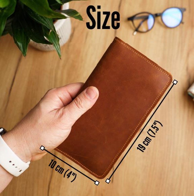May include: A brown leather wallet, measuring 10 cm wide and 19 cm tall, is held in a hand. The wallet has a stitched border and a smooth surface. The word "Size" is visible in the image.