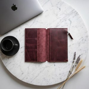 Può includere: Un portafoglio o organizer in pelle color bordeaux scuro aperto, dotato di molteplici scomparti per carte sul lato sinistro e uno scomparto con cerniera sul lato destro con una linguetta in pelle abbinata, con finitura liscia e opaca.