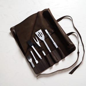 May include: A brown leather barbecue tool roll with stainless steel utensils. The set includes a spatula, fork, basting brush, and tongs. The roll is open, displaying the tools, and has a strap for carrying. The background is a white wooden surface.