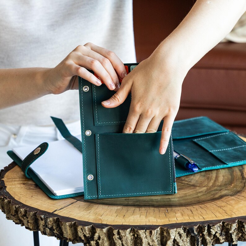 K&ouml;nnte beinhalten: Ein teal-farbenes Leder-Journal und Organizer-Set. Das Journal ist ge&ouml;ffnet und zeigt leere Seiten, w&auml;hrend der Organizer mehrere Taschen und Druckkn&ouml;pfe aufweist. Das Set wird auf einem Holztisch pr&auml;sentiert.