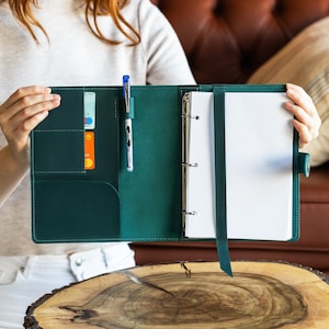 Könnte beinhalten: Offener, dunkelgrüner Lederordner mit Stift, Kreditkarten und weißem Papier. Der Ordner hat einen Drei-Ring-Mechanismus und einen Riemen, um die Seiten an Ort und Stelle zu halten. Der Ordner wird von einer Person offen gehalten.
