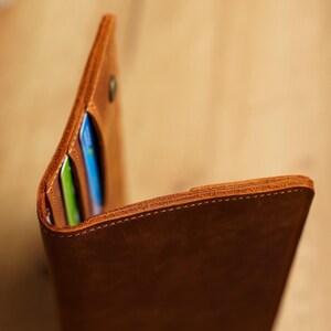 May include: A brown leather wallet, partially open to reveal card slots. The wallet has a button closure and visible stitching along the edges. The leather has a textured appearance. The wallet contains colorful cards.