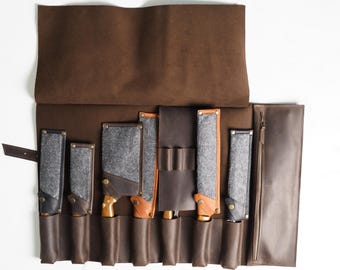 May include: A brown leather knife roll with multiple knife sheaths. The sheaths are made of gray felt and brown leather, with wooden handles visible. The roll is open, displaying the knives and their protective covers.