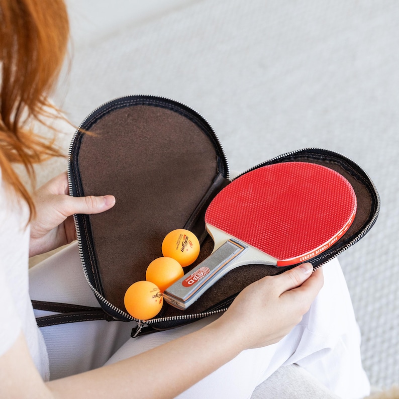 K&ouml;nnte beinhalten: Ein roter und schwarzer Tischtennisschl&auml;ger mit wei&szlig;em Griff und drei orangefarbenen Tischtennisb&auml;llen befinden sich in einem braunen Rei&szlig;verschluss-Etui. Das Etui ist ge&ouml;ffnet und zeigt den Inhalt.