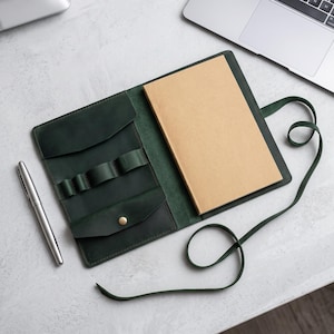 Könnte beinhalten: Ein dunkelgrüner Notizbuchumschlag aus Leder mit integriertem Stiftehalter, einer kleinen Tasche mit Messingdruckknopf und einem langen Lederband zum Verschließen. Ein beigefarbenes Notizbuch liegt darin und ein silberner Stift liegt daneben.