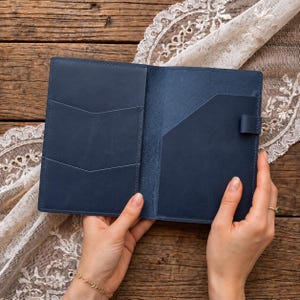 Pode incluir: Uma capa de caderno em couro azul-marinho com dois bolsos sobrepostos em forma de V no lado esquerdo e um bolso grande com um suporte para caneta no lado direito. O interior é feito de couro liso com um acabamento fosco.