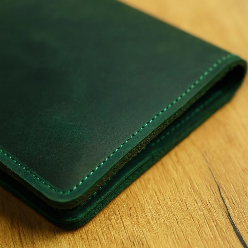 May include: Close-up of a dark green leather wallet. The wallet is folded, revealing the edges and stitching. The stitching is a lighter green colour. The leather has a slightly distressed appearance. The wallet is on a wooden surface.