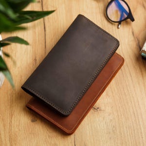 May include: Two leather wallets, one dark brown and one tan, are stacked on a wooden surface. The wallets are rectangular with rounded corners and stitched edges. The dark brown wallet is on top, partially obscuring the tan one.