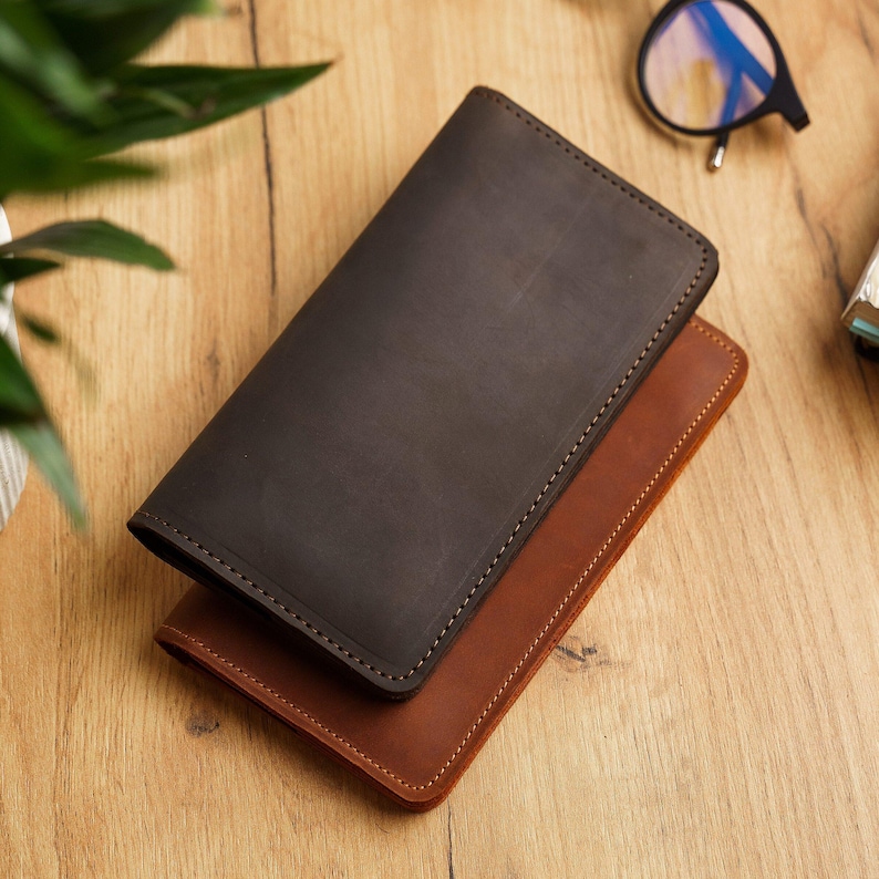 May include: Two leather wallets, one dark brown and one tan, are stacked on a wooden surface. The wallets are rectangular with rounded corners and stitched edges. The dark brown wallet is on top, partially obscuring the tan one.