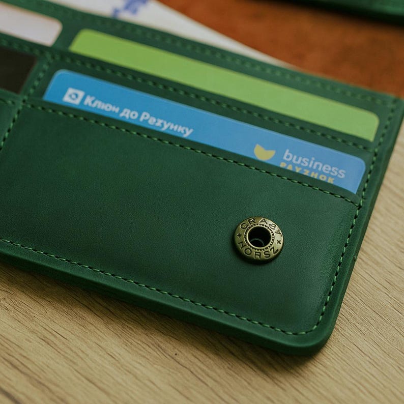 May include: A green leather wallet with card slots, open to reveal cards with Ukrainian and English text. The wallet has a metal snap closure. The stitching is visible.