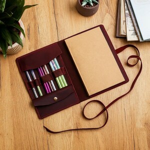 Könnte beinhalten: Geöffnete burgunderfarbene Stricknadel-Tasche aus Leder mit einem beigefarbenen Notizbuch und verschiedenen farbigen Nadeln. Die Tasche hat einen Druckknopfverschluss und einen langen Riemen. Die Nadeln sind in verschiedenen Größen und Farben.