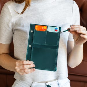 May include: Teal leather wallet with multiple card slots and a zipped compartment. The wallet is held open, revealing credit cards inside. The stitching is white, and the wallet has a small wrist strap.