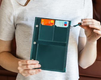 May include: Teal leather wallet with multiple card slots and a zippered compartment. The wallet is held open, revealing credit cards inside. The stitching is white, and the wallet has a small wrist strap.